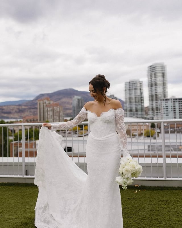 Bride holding the train of a fitted off-the-shoulder lace wedding gown